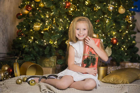 Happy cute girl sit near christmas tree with red gift box. New year holiday, female child with present in festive decorated home room.の写真素材
