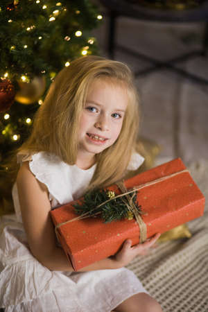 Happy cute girl sit near christmas tree with red gift box. New year holiday, female child with present in festive decorated home room.の写真素材