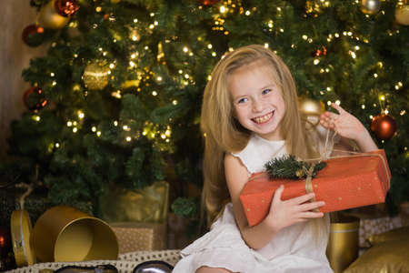 Happy cute girl sit near christmas tree with red gift box. New year holiday, female child with present in festive decorated home room.の写真素材