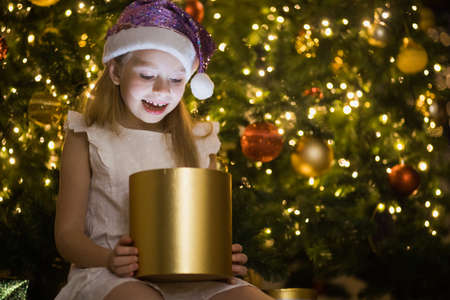 Little girl sits in decorated christmas room opening magic gift box with light in it. Happy child in Santa glitter hat near New Year fir tree.の写真素材