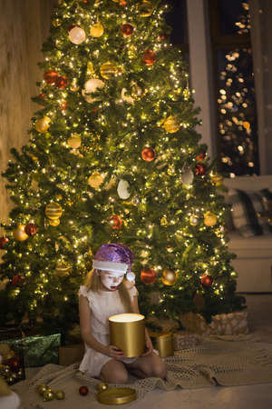 Little girl sits in decorated christmas room opening magic gift box with light in it. Happy child in Santa glitter hat near New Year fir tree.の写真素材