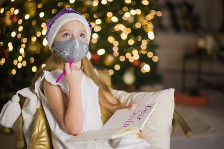 Cute little girl in facial mask writing letter to Santa Claus near christmas tree. Female child celebrate new year during coronavirus lockdown. Quarantine xmas concept.の写真素材