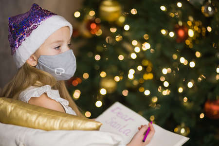 Cute little girl in facial mask writing letter to Santa Claus near christmas tree. Female child celebrate new year during coronavirus lockdown. Quarantine xmas concept.の写真素材