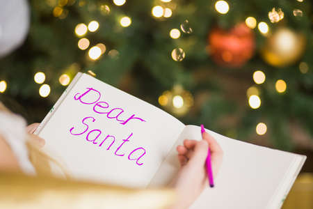 Happy little girl writing letter to Santa in decorated christmas room. Child smile and wish gifts near festive xmas fir-tree. New Year and winter holidays concept.の写真素材