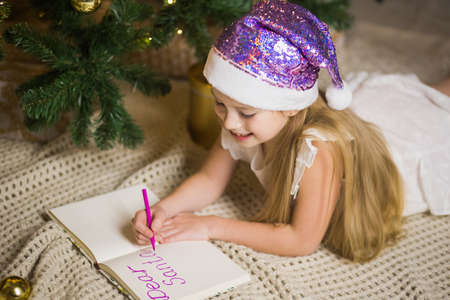 Happy little girl writing letter to Santa in decorated christmas room. Child smile and wish gifts near festive xmas fir-tree. New Year and winter holidays concept.の写真素材