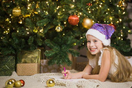 Happy little girl writing letter to Santa in decorated christmas room. Child smile and wish gifts near festive xmas fir-tree. New Year and winter holidays concept.の写真素材