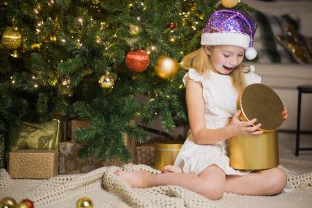 Little girl sits in decorated christmas room opening magic gift box with light in it. Happy child in Santa glitter hat near New Year fir tree.の写真素材