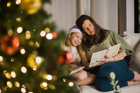 Mother and daughter read book with Christmas tree lights on. New year, family and love concept.の写真素材