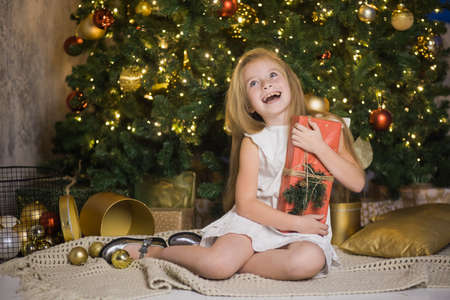 Happy cute girl sit near christmas tree with red gift box. New year holiday, female child with present in festive decorated home room.の写真素材