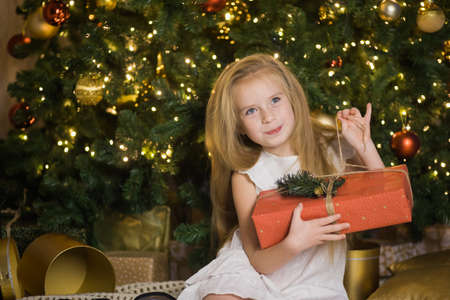 Happy cute girl sit near christmas tree with red gift box. New year holiday, female child with present in festive decorated home room.の写真素材
