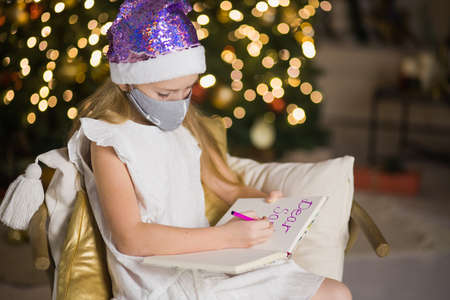 Cute little girl in facial mask writing letter to Santa Claus near christmas tree. Female child celebrate new year during coronavirus lockdown. Quarantine xmas concept.の写真素材