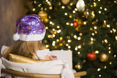 Cute little girl in facial mask writing letter to Santa Claus near christmas tree. Female child celebrate new year during coronavirus lockdown. Quarantine xmas concept.の写真素材