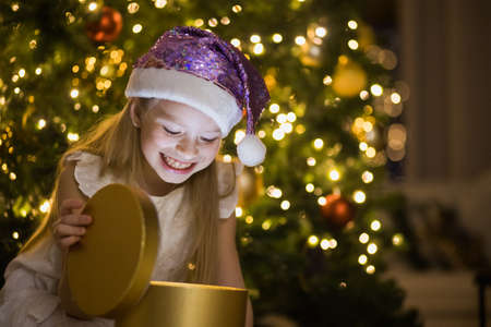 Little girl sits in decorated christmas room opening magic gift box with light in it. Happy child in Santa glitter hat near New Year fir tree.の写真素材