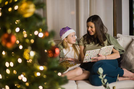 Mother and daughter read book with Christmas tree lights on. New year, family and love concept.の写真素材