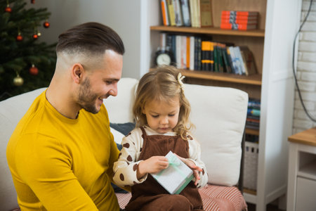 Young father give gift box to little toddler daughter on Christmas. Man and little girl sit on couch together near decorated fir tree.の写真素材