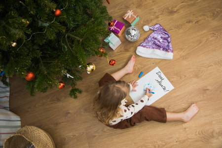 Little toddler girl sit on floor and write or draw letter for santa. Cute kid make wish on Christmas time at home.の写真素材