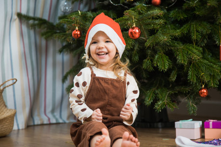 Cute little girl with disco ball sit on floor neat Christmas tree. Toddler female child in santa hat wait for new year holiday.の写真素材