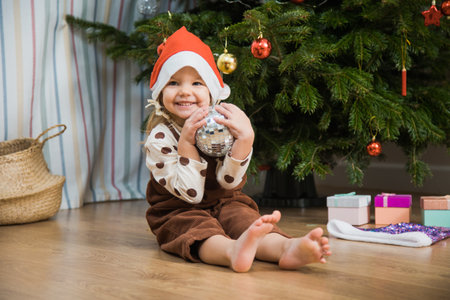 Cute little girl with disco ball sit on floor neat Christmas tree. Toddler female child in santa hat wait for new year holiday.の写真素材