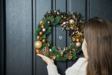 Young woman hanging christmas festive wreath on house door. Traditional home decoration on winter holidays, female hands holding fir tree handmade wreath on doorway.の写真素材