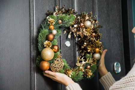 Young woman hanging christmas festive wreath on house door. Traditional home decoration on winter holidays, female hands closeup holding fir tree handmade wreath on doorway.の写真素材