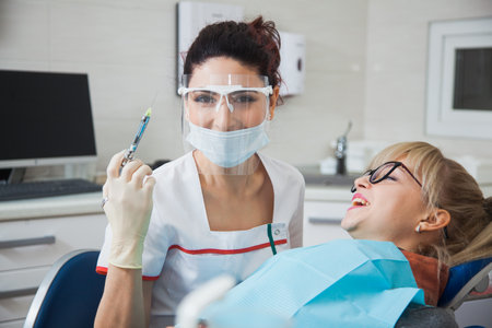 Female dentist in protection mask holding syringe with anesthetic. Stomatologist and patient at clinic office. Toothcare conceptの写真素材