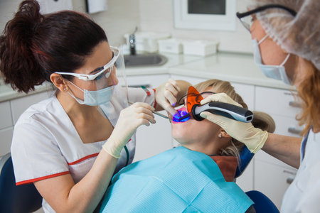 Closeup shot of dentist and assistant in protection mask working with tooth cavity and plague. Patient repair aching teeth. Stomatologist and patient at clinic office. Toothcare conceptの写真素材