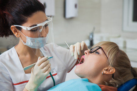 Closeup shot of dentist in protection mask working with tooth cavity and plague. Patient on stomatology chair repair aching teeth. Stomatologist and patient at clinic office. Toothcare conceptの写真素材