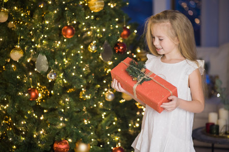 Happy cute girl sit near christmas tree with red gift box. New year holiday, female child with present in festive decorated home room.の写真素材