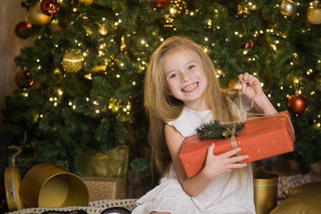Happy cute girl sit near christmas tree with red gift box. New year holiday, female child with present in festive decorated home room.の写真素材