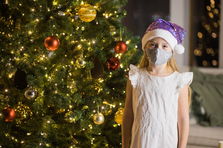 Little girl in protective mask and santas hat in decorated christmas room. New Years eve staying at home during covid lockdown.の写真素材