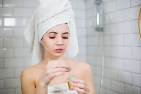 Young woman in white towel apply moisturising oil on face skin. Self care, beauty and skin care concept.の写真素材