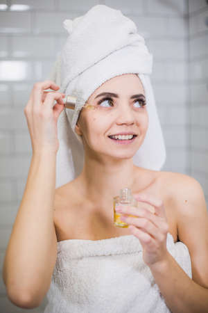 Young woman in white towel apply moisturising oil on face skin. Self care, beauty and skin care concept.の写真素材