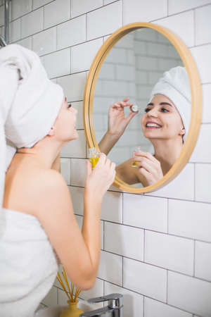 Young woman in white towel apply moisturising oil on face skin. Self care, beauty and skin care concept.の写真素材