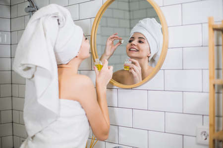Young woman in white towel apply moisturising oil on face skin. Self care, beauty and skin care concept.の写真素材