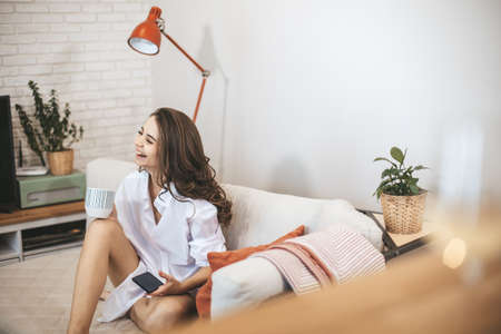 Young woman using smartphone sitting on couch at home. Young person drinks coffe in big mug and serfing internetの写真素材