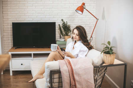 Young woman using smartphone sitting on couch at home. Young person drinks coffe in big mug and serfing internetの写真素材