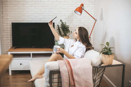 Young woman makes selfie sitting on couch at home. Young person drinks coffe in big mug and use smartphoneの写真素材
