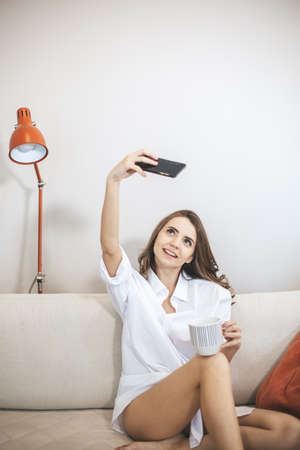 Young woman makes selfie sitting on couch at home. Young person drinks coffe in big mug and use smartphoneの写真素材