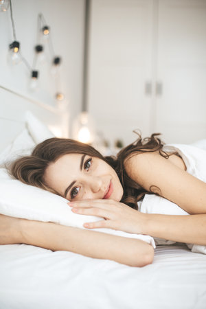 Beautiful young woman waking up in comfortable bed on white fresh sheets.の写真素材