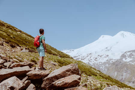 Man with red backpack stand on rocky mountain and look at snowy peak of Elbrus.の写真素材