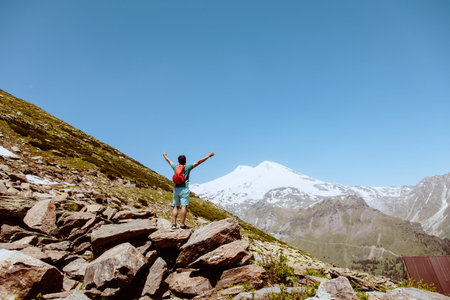 Man with red backpack stand on rocky mountain and look at snowy peak of Elbrus.