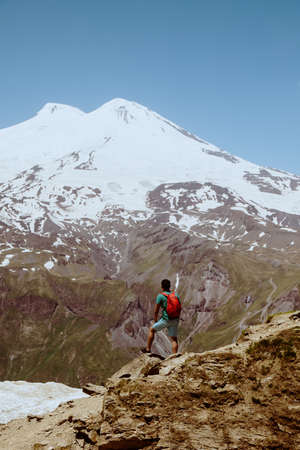 Man with red backpack stand on rocky mountain and look at snowy peak of Elbrus.の写真素材