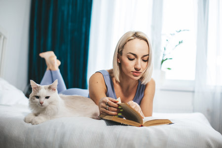 A beautiful woman reads a book in her bedroom with her cat by her side, female pet lover.の写真素材