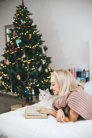 A blonde-haired woman is reading book next to a cat during Christmas with decorated fir treeの写真素材