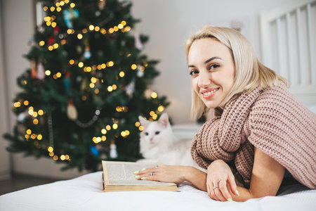 A blonde-haired woman is reading book next to a cat during Christmas with decorated fir treeの写真素材