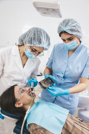 Patient in a dental chair with an open mouth receives treatment from the dentist while an assistant captures the process on a smartphoneの写真素材