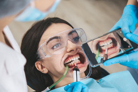 Patient in a dental chair with an open mouth receives treatment from the dentist while an assistant captures the process on a smartphoneの写真素材