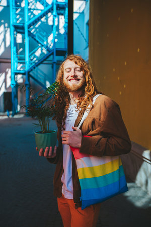 A man with long curly red hair is holding a plant in a green pot. He is smiling and he is happy