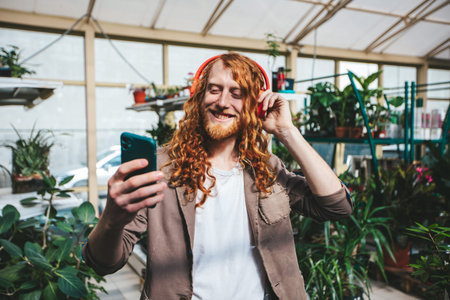 A man with red hair is holding a cell phone and wearing headphones. He is smiling and he is enjoying...