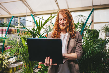Young business owner is smiling while using a laptop to manage his plant nursery. He is standing in the middle of his shop surrounded by lush greeneryの写真素材
